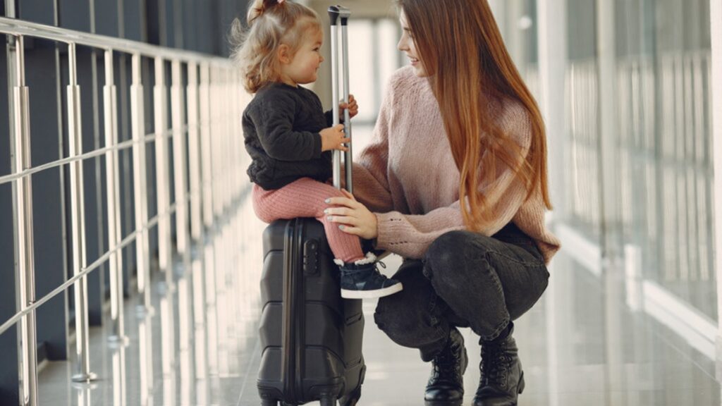 A mother kneels beside her young daughter, who sits on a suitcase in an airport