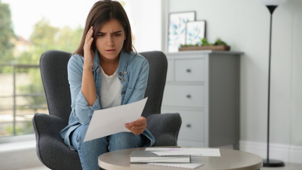 Worried young woman in a denim shirt reading a letter in a gray armchair at home